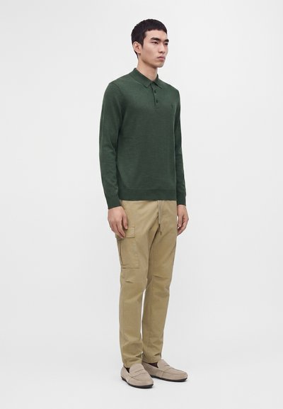 Homme portant un polo vert foncé à manches longues, un pantalon cargo beige et des chaussures beige clair à enfiler, debout devant un fond uni.