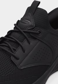 Κοντινό πλάνο ενός μαύρου αθλητικού παπουτσιού με ανάγλυφο δίχτυ, δερμάτινες επιφάνειες, στρογγυλά κορδόνια και ρυθμιστικό στο κολάρο με ανάγλυφο λογότυπο "S".