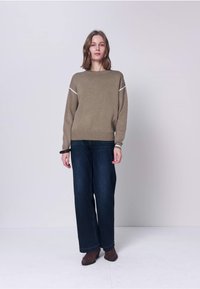 Femme debout portant un pull marron avec des rayures blanches, un jean large foncé et des bottines marron, sur un fond uni clair.