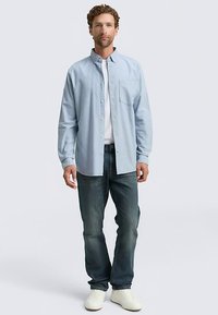 Hellblaues Hemd mit Knöpfen und einer Brusttasche, getragen über einem weißen T-Shirt. Kombiniert mit dunkelblauen Jeans und weißen Turnschuhen.