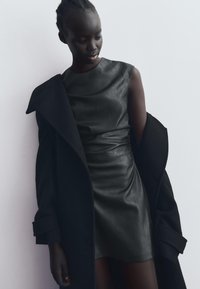 Rochie din piele neagră cu o siluetă ajustată, având un guler înalt și frunză subtilă. Purcată cu o haină neagră din țesătură texturată.