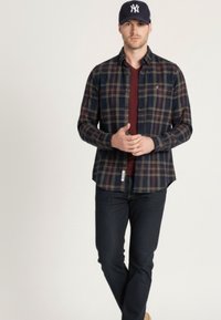 Camisa de manga larga de cuadros en colores azul marino, verde y burdeos, usada sobre una camiseta de cuello en V burdeos, combinada con vaqueros oscuros y una gorra.