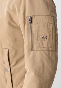 Kaki jacka med mjuk textur, ficka med dragkedja på överarmen och ett runt emblem med initialer på ärmen.