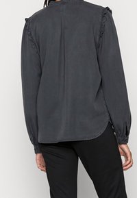 Blusa grigia scura a maniche lunghe con dettagli arricciati sulle spalle e polsini con bottone. Presenta un scollo rotondo e un orlo leggermente curvato.