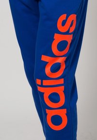 Modré tepláky vyrobené z měkké látky, na boku s velkým oranžovým logem "adidas". Design je jednoduchý s rovnými nohavicemi.