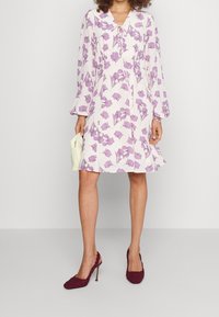 Weißes Kleid mit lila Blättern-Muster, langen Ärmeln und Bindeband am Ausschnitt. Kombiniert mit burgunderfarbenen High Heels und einer kremfarbigen Clutch.