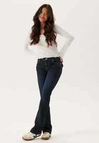 Dopasowane ciemnoniebieskie dżinsy z rozszerzanym krojem nogawki, wykonane z denimu o gładkiej fakturze. Połączone z białą bluzką z długim rękawem.