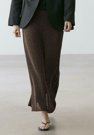 Osoba spacerująca w długiej brązowej dzianinowej spódnicy, czarnym blezerze i czarnych sandałach na płaskiej podeszwie, na jasnej podłodze.