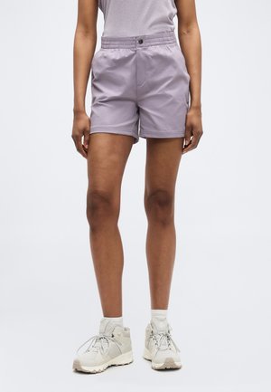 Persoană purtând pantaloni scurți cu talie elastică lila, top fără mâneci gri, șosete albe și pantofi de drumeție gri deschis, stând în fața unui fundal simplu.