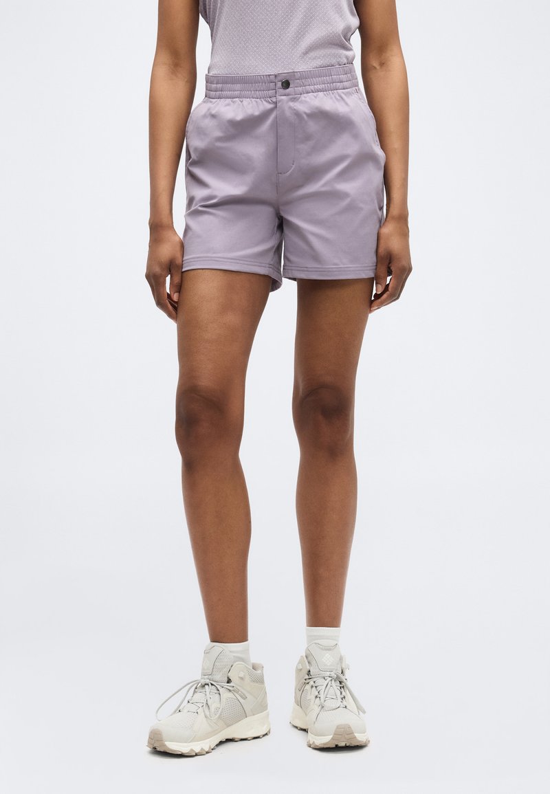 Άτομο που φοράει λιλά σορτς με ελαστική μέση, γκρι αμάνικο τοπ, λευκές κάλτσες και ανοιχτό γκρι παπούτσια πεζοπορίας, στέκεται μπροστά σε ομοιόμορφο φόντο.