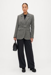 Blazer cinza em padrão pied-de-poule com cinto, dois bolsos frontais e lapelas. Combinado com calças largas pretas e sapatos de plataforma pretos robustos.