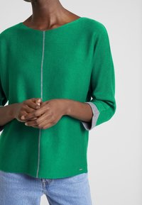 Groene gebreide top met een bootnek en driekwartmouwen; heeft een grijze accent aan de manchetten en een verticale grijze lijn aan de voorkant.