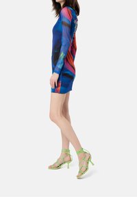 Vestido mini de manga larga, ajustado y colorido, con un diseño abstracto en azul, rojo y verde. También presenta sandalias de tacón alto verdes con tiras.