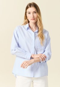 Camicia azzurro chiaro a maniche lunghe, con bottoni e sottili righe verticali bianche, colletto, dotata di taschino sul petto e orlo arrotondato.