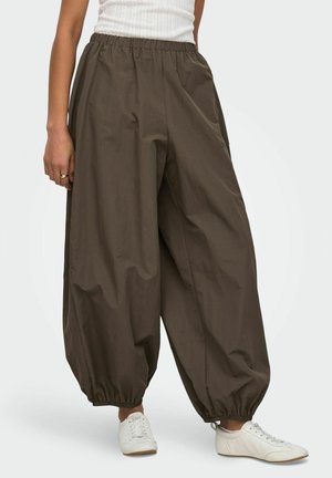 Osoba ubrana w luźne, ciemnobrązowe spodnie z elastyczną talią i zebranymi nogawkami, biały bezrękawnik oraz białe trampki stoi na jasnym tle.