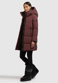 Burgundy pufi kabát kapucnival, oldalsó cipzáras zsebekkel és fekete elülső cipzárral. A modellen fekete leggings és fényes fekete hócipő van.