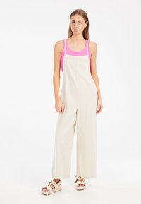 Overoles de algodón beige con piernas anchas y tirantes, combinados con una camiseta de tirantes rosa brillante y sandalias beige claras.