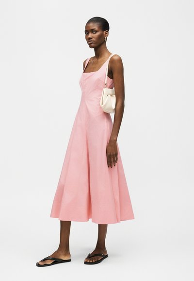 Μια γυναίκα στέκεται φορώντας ένα ροζ αμάνικο μίντι φόρεμα, μαύρες σαγιονάρες, μικρή λευκή τσάντα ώμου και χρυσούς κρίκους σκουλαρίκια, πάνω σε ένα απλό φόντο.