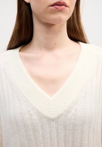 Sweter w serek w miękkim, kremowym splocie. Posiada prążkowaną teksturę i otwarte dziewiarskie detale, z luźnym krojem w okolicach szyi i ramion.