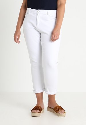 Vrouw draagt witte opgerolde enkelbroeken, een zwart topje en bruine platform-slippers, staand op een witte vloer.