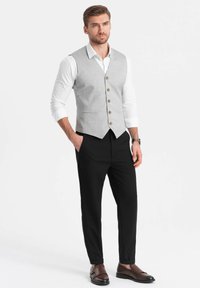 Gilet grigio chiaro con una forma aderente, cinque bottoni marroni, abbinato a pantaloni neri e scarpe marroni, con una texture liscia.