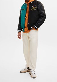 Veste universitaire noire et verte, sweat à capuche orange, pantalon de survêtement crème et baskets grises avec des accents blancs ; comprend des logos brodés et des poignets rayés.