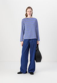 Long-sleeve striped top in blue and white, paired with wide-leg blue jeans, holding a black shoulder bag, standing on a plain background.