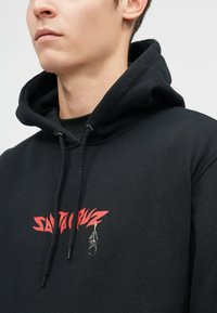 Μαύρη κουκούλα με κόκκινη εκτύπωση γραφικών και μια μικρή απεικόνιση μπροστά. Φτιαγμένη από μαλακό ύφασμα με ρυθμιζόμενο κορδόνι.