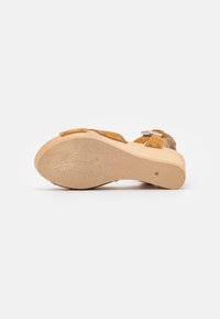Bruine suède sandaal met een lichte houten plateauzool, voorzien van metalen gespen en een gestructureerde grip voor tractie.