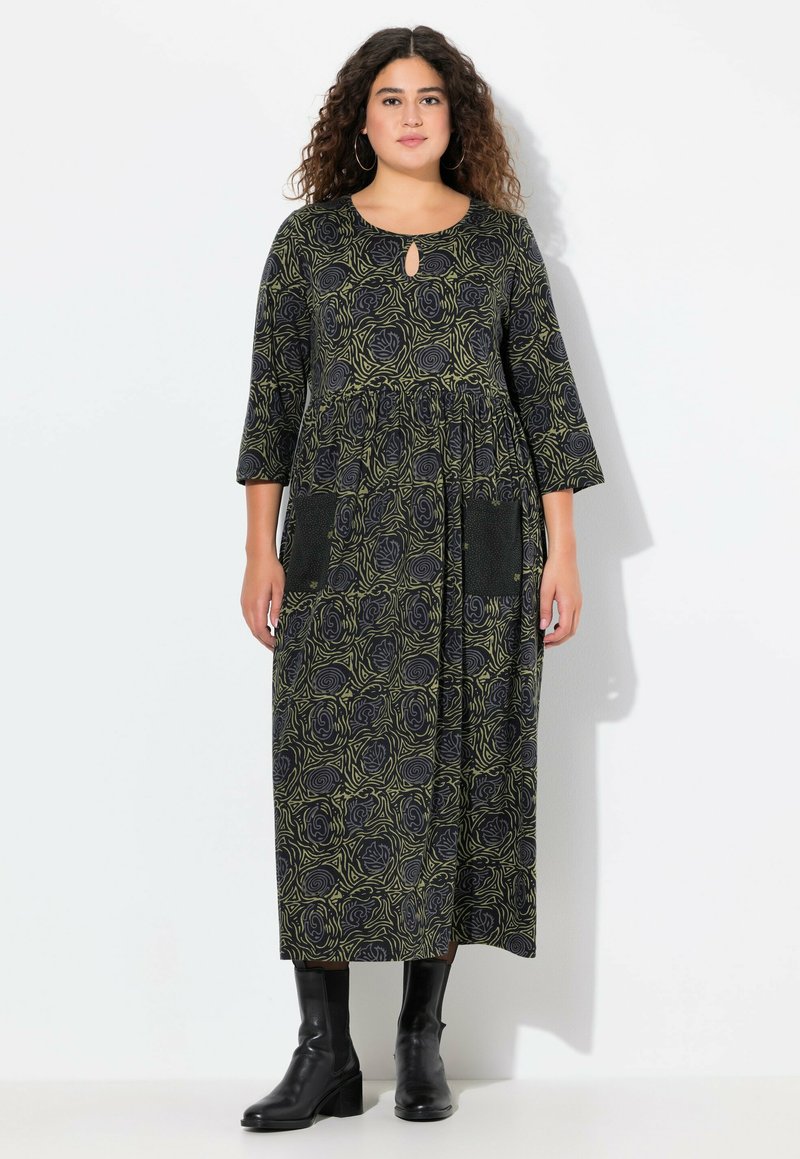 Černé dlouhé šaty s zeleným vzorem růží, kulatým výstřihem, detailem ve tvaru klíče, třičtvrtečními rukávy a dvěma předními kapsami. Nosené s černými kotníkovými botami.