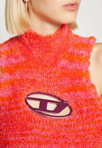 Haut sans manches duveteux aux couleurs vives orange et rose. Présente un détail logo découpé et un design à col montant. Texture douce.