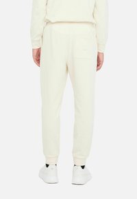 Joggers de algodón en crema claro con un corte recto, puños acanalados y un bolsillo trasero. Combinados con zapatillas blancas, mostrando un diseño relajado.