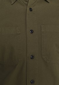 Olivgrünes Hemd aus strukturiertem Stoff mit spitzem Kragen, schwarzen Knöpfen und zwei Brusttaschen mit Klappen.