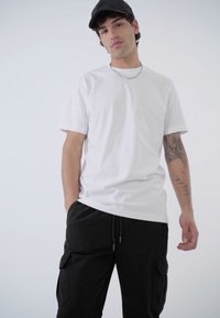 T-shirt en coton blanc à manches courtes, associé à un pantalon cargo noir avec des poches latérales, porté avec une casquette noire et un collier en chaîne.