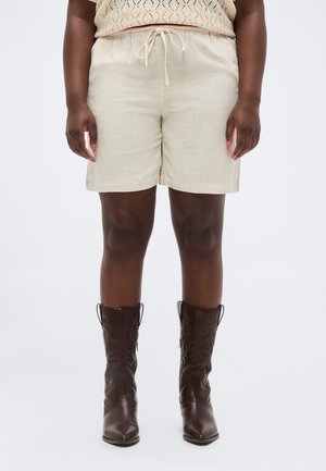 Személy, aki bézs színű, zsinórral szabályozható rövidnadrágot, sötétbarna, középmagas szárú bőr csizmát, valamint deréktól lefelé látható bézs színű kötött felsőt visel.