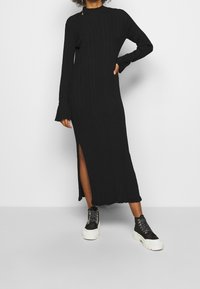 Vestito nero a costine con maniche lunghe, scollatura alta e spacco laterale. Abbinato a sneakers alte nere con suole bianche.