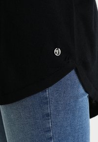 Musta neulepaita, jossa on pyöreä helma ja pieni hopeinen logo. Yhdistetty sinisiin farkkuihin, joissa sileä pinta.