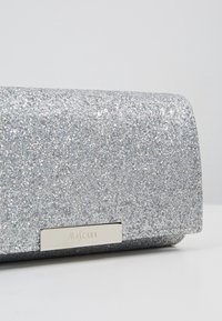 Pochette rectangulaire argentée pailletée avec une petite plaque métallique gravée du mot « Mascara » à l'avant.