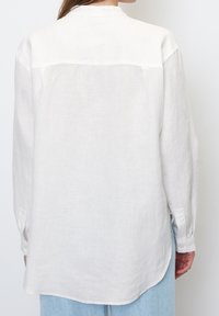 Weißes Leinenshirt mit einem Button-Down-Kragen, langen Ärmeln und einem geschwungenen Saum. Glatte Textur mit lockerem Sitz und dezenten Nahtdetails.