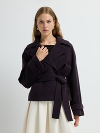Cappotto viola avvolgente con ampi revers e cintura, realizzato in tessuto morbido e maniche spaziose, abbinato a una leggera gonna plissettata.