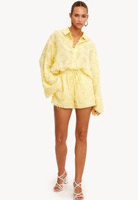 Mujer vestida con una camisa de botón amarilla de manga larga texturizada y pantalones cortos a juego, con tacones altos blancos con tiras, de pie contra un fondo simple.