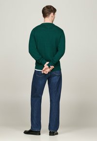 Maglione lavorato a maglia verde scuro con maniche lunghe, indossato sopra jeans in denim blu. Il maglione presenta un collo a girocollo e polsini a coste.