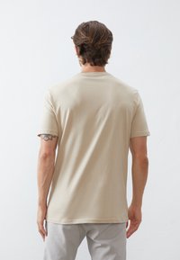 Camiseta de algodón beige con mangas cortas, cuello redondo y textura suave, mostrada por la parte de atrás sobre un fondo blanco liso.