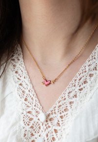 Guld kæde med en pink hjertevedhæng med pil-accenter. Vedhænget har en blank finish mod en blonderbluse.
