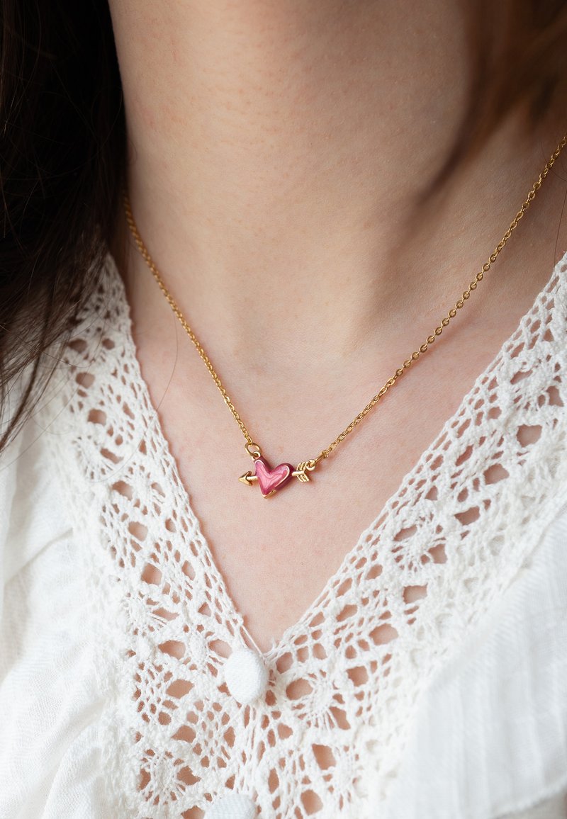 Guld kæde med en pink hjertevedhæng med pil-accenter. Vedhænget har en blank finish mod en blonderbluse.