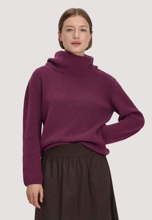 Vrouw met kort haar die een paarse geribbelde coltrui en een donkerbruine rok met een elastische tailleband draagt, staat met één hand op haar heup.