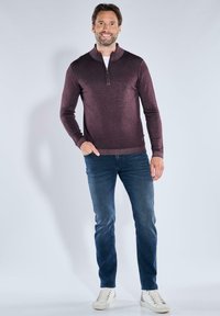 Bordeauxrotem Reißverschluss-Pullover mit hohem Kragen, kombiniert mit blauen Jeans und weißen Turnschuhen. Die Pulloverstruktur wirkt weich und körperbetont.