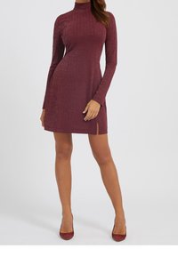 Femme portant une robe bordeaux côtelée à manches longues et col haut, avec une petite fente à l'avant, accompagnée de talons assortis à bout pointu.