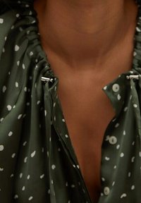 Blusa verde con scollatura arricciata, caratterizzata da pois bianchi e dettagli in metallo argentato. Tessuto morbido con un design drappeggiato.