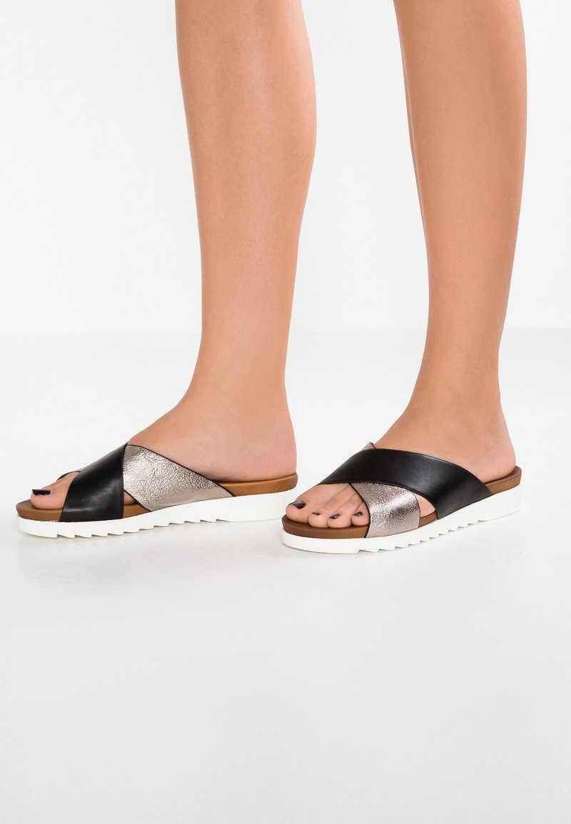 Schwarze und metallische Rutschsandalen mit einem Überkreuzdesign, weißer strukturierten Sohle und offener Zehenpartie, die einen eleganten und modernen Look präsentieren.